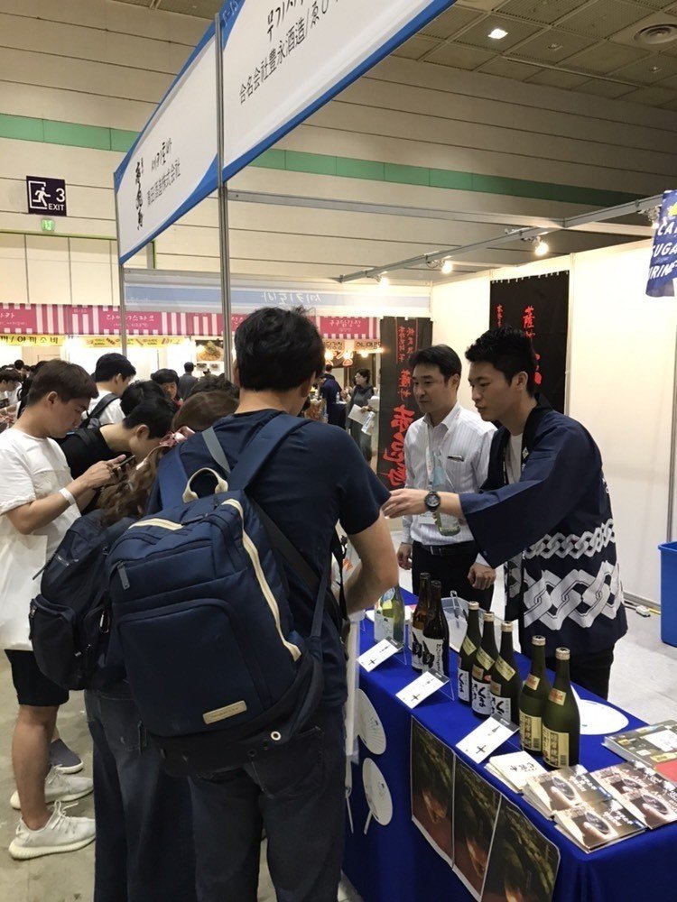 韓国の焼酎イベントのブース。はっぴ姿の学生時代の橋本が来場者に焼酎を勧め、その左奥にゑびす酒造の田中社長が立つ。