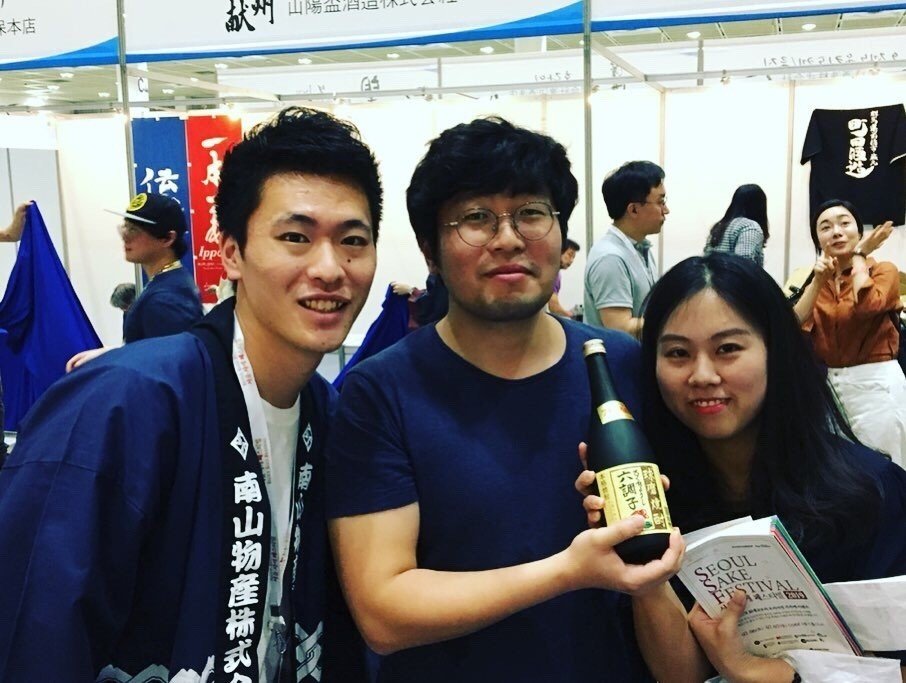 イベント会場で、はっぴ姿の学生時代の橋本と、六調子酒造の焼酎を手にした来場者2人。